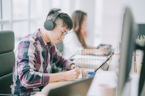 joven chino asiático escuchando con auriculares, escribiendo en el bloc de notas e aprendiendo usando una tableta digital en su lugar de trabajo photo