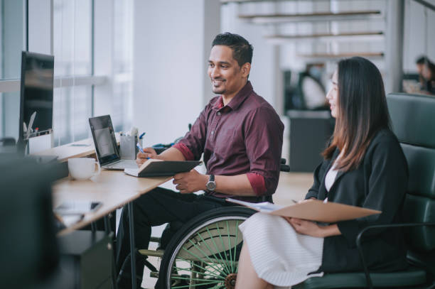 operaio indiano con colletto bianco su sedia a rotelle che ha un'allegra conversazione di discussione con la sua collega cinese asain che lavora in una postazione di lavoro creativa accanto alla finestra - working physical impairment people occupation foto e immagini stock