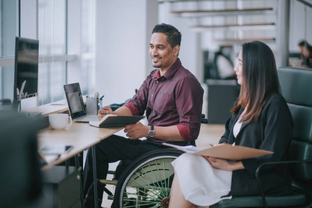 indian white collar male worker in wheelchair having cheerful discussion conversation with his female asain chinese colleague coworking in creative office workstation beside window - 傷殘人士設施 圖片 個照片及圖片檔