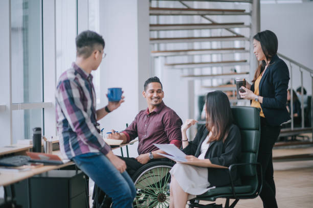 col blanc indien travailleur masculin en fauteuil roulant ayant une discussion joyeuse menant la conversation avec un collègue dans un poste de travail de bureau créatif à côté de la fenêtre - job photos et images de collection