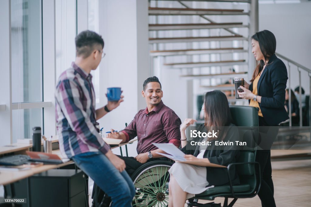 Indian white collar male worker in wheelchair having cheerful discussion leading conversation with colleague in creative office workstation beside window Diversity Stock Photo