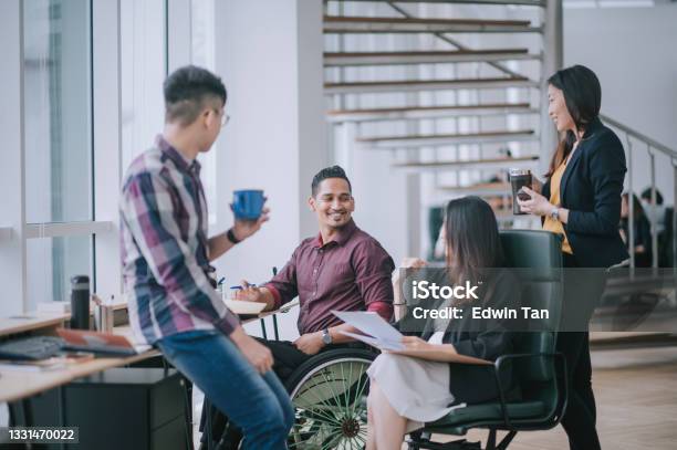 Indian White Collar Male Worker In Wheelchair Having Cheerful Discussion Leading Conversation With Colleague In Creative Office Workstation Beside Window Stock Photo - Download Image Now