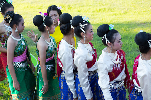 Thai teacher is instructing young traditional period dressed female dancers standing in a row. Women are having flowers on hair. Scene is at history event at old ruined temple. Group is exercising for evening event. 2019 was historical event at and around temple ruin Wat Wihan Thong Historical Site with food market and dance shows events for publicity and admission free. Typical thai true classic market and event festival