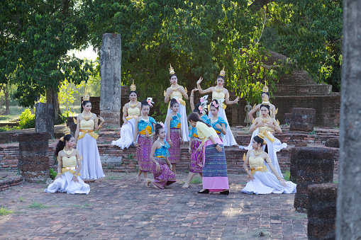 Young female rum thai dancers are exercising with teacher at ruins of temple in Phitsanulok near river. Dancers are traditionally dressed. Some have flower decor and others have golden crowns. 2019 was historical event at and around temple ruin Wat Wihan Thong Historical Site with food market and dance shows events for publicity and admission free. Typical thai true classic market and event festival