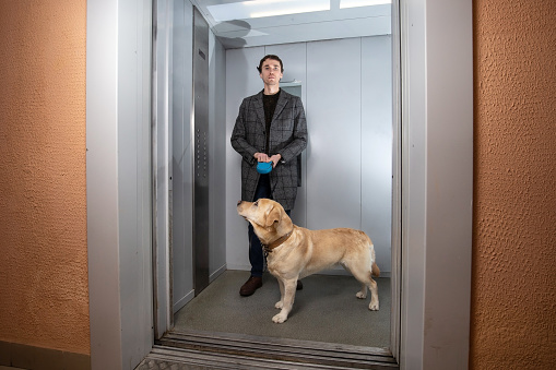 Handsome stylish man in a checked coat standing with labrador dog in elevator and looking at camera.