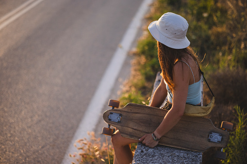 High angle view of a young unrecognizable Caucasian woman having fun on her own, sitting on the sideroad wall with her longboard and enjoying the sunset