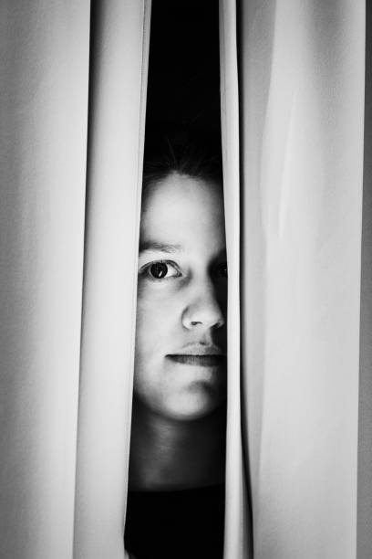 Wary young woman peers cautiously through her curtains Suspicious, anxious young woman looking through a window. woman stealth stock pictures, royalty-free photos & images