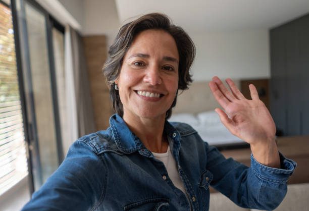 Happy woman at home greeting on a video call Happy Latin American woman at home greeting on a video call and smiling while holding their cell phone - lifestyle concepts selfie stock pictures, royalty-free photos & images
