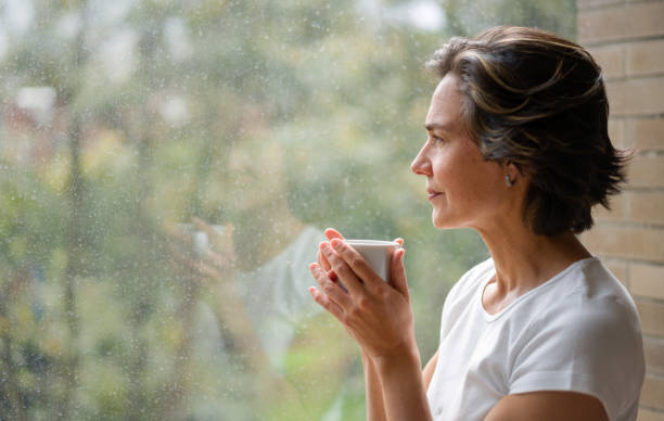 mujer bebiendo una taza de café mientras mira por la ventana - 45 49 años fotografías e imágenes de stock