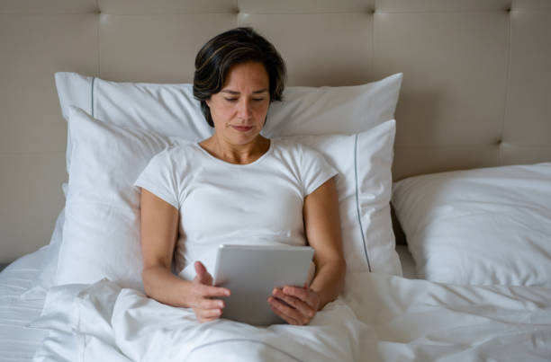 woman relaxing in bed and reading a book on her e-reader - kindle e reader book reading imagens e fotografias de stock