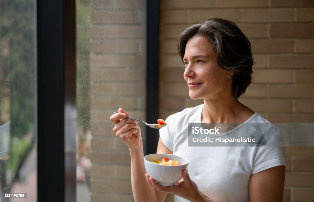 Happy woman at home eating a healthy breakfast while looking out the window Happy woman at home eating a healthy breakfast while looking out the window - domestic life concept Eating Stock Photo