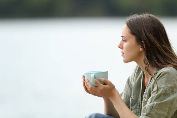 femme tenant une tasse de café contemplant un lac - at the beach photos et images de collection