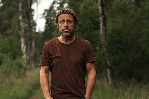 Blonde man with a stubble beard in a brown t-shirt on a forest path in the summer. Front view.