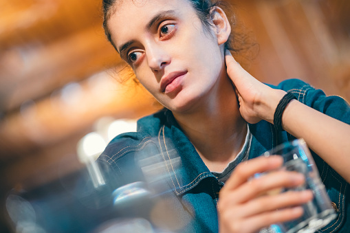 In this indoor image with copy space, an Asian/Indian young woman takes a break and contemplates holding a glass of water in a restaurant. She is in a casual dress.