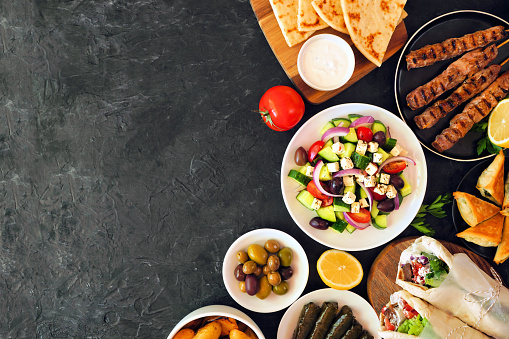 Greek food side border, top view on a dark background. Souvlaki, gyros wraps, salad, spanakopita, dolmades, pita and tzatziki. Copy space.