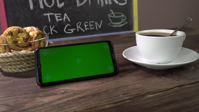Hot drink, cookies and smartphone on the table in the cafe.Smartphone green screen.