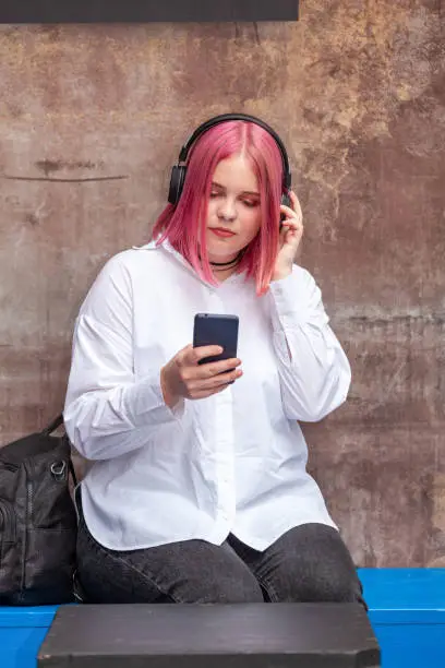 Outdoors portrait of 22 year old woman in white shirt with pink hair wearing headphones listening to music