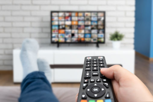 Man watching TV, lying on sofa, legs on table. Person holding remote control in living room