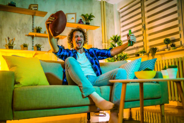 Young happy man watching American football on TV Excited young happy man watching American football on TV at night football league stock pictures, royalty-free photos & images