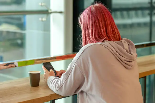 Indoors portrait of 20 year old woman in hooded shirt with pink hair holding mobile phone