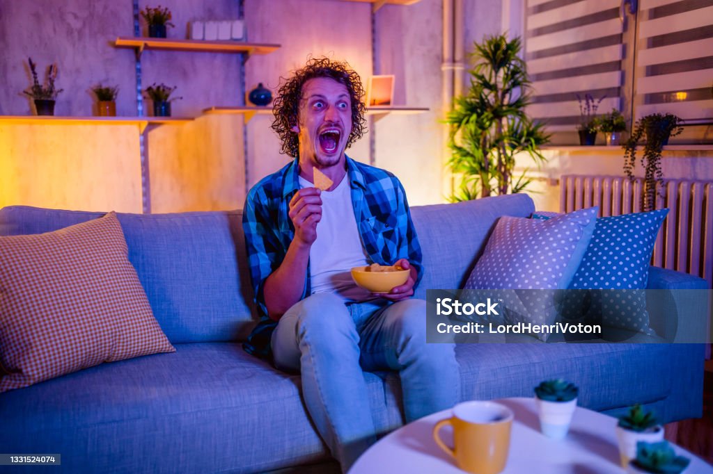 Man scared by horror movie Young scared man watching horror movie and eating snacks in the living room at night Television Set Stock Photo