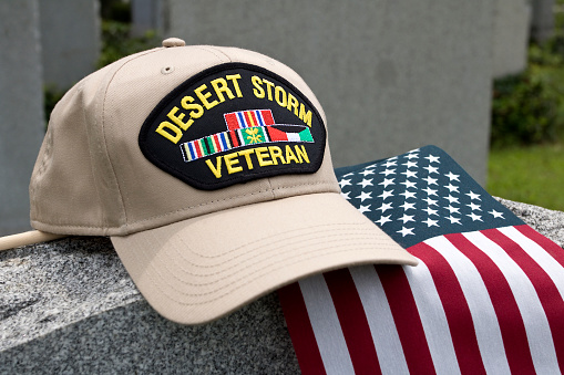 Desert Storm Veterans cap inscribed with text depicting the Gulf War from January 17, 1991 – February 28, 1991, sitting on tombstone with American Flag.