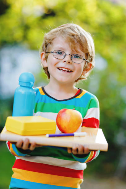 menino feliz da pré-escola com livros, maçã e garrafa de bebida em seu primeiro dia no ensino fundamental ou berçário. criança sorridente, estudante de óculos, ao ar livre. de volta ao conceito de educação escolar. - water child bottle little boys - fotografias e filmes do acervo