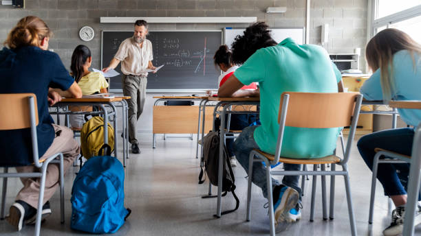 Mature caucasian man teacher hands out exams to multiracial high school students. Students ready to take exam. Mature caucasian man teacher hands out exams to multiracial high school students. Students ready to take exam. Education concept. high school stock pictures, royalty-free photos & images