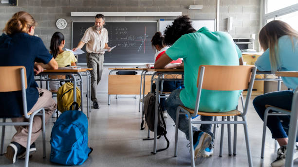 mature caucasian man teacher hands out exams to multiracial high school students. students ready to take exam. - high school imagens e fotografias de stock