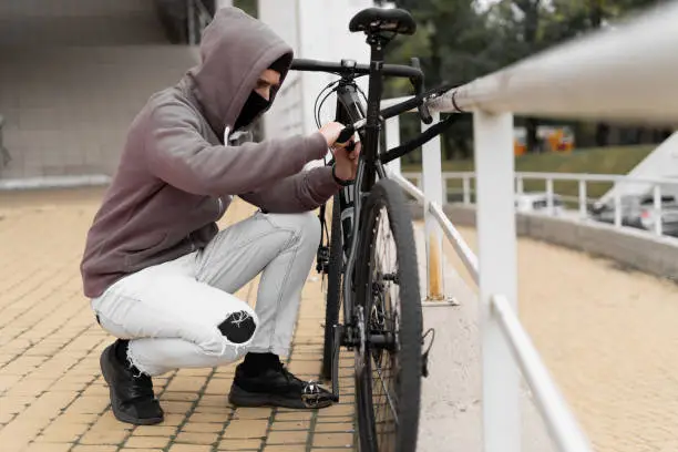 A thief in a balaclava and a hood breaks the lock on a bicycle in the street during the day. Closed face and hacking. Stealing bicycles. Place for your text.