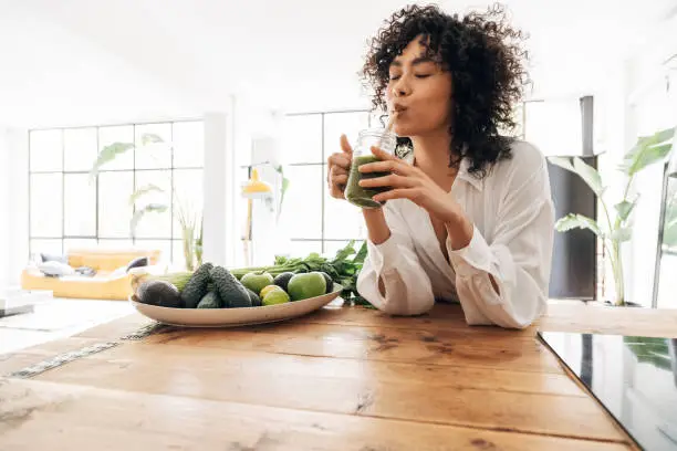 Photo of Young african american woman drinking green juice with reusable bamboo straw in loft apartment. Copy space