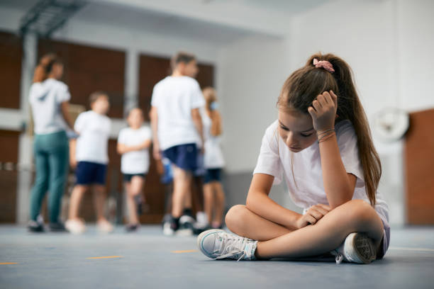 Sad schoolgirl feeling left out during physical activity class at school gym. Elementary student sitting away from her classmates and teacher and feeling sad during physical education class. physical education stock pictures, royalty-free photos & images