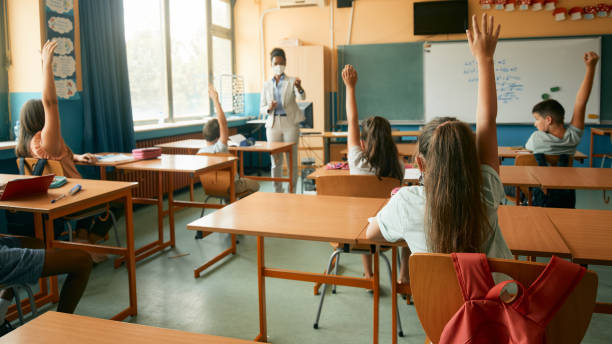istock Back view of school children raising their hands to answer teacher's question during a class in the classroom. 1331595043