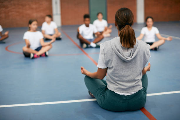 Rear view of sports teacher practicing Yoga with her students at school gym. Back view of PE teacher leading Yoga class and meditating with elementary students at school gym. mindfulness kids stock pictures, royalty-free photos & images