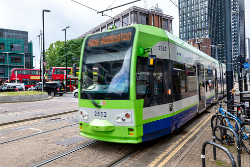 Croydon, England - July 30, 2021: A tram bound for Beckenham Junction in Kent, about to leave East Croydon tram station in Surrey. Both places are in the suburban Greater London area. The building on the right is affectionately known as The Threepenny Bit Building because of its unusual shape and is a relic of the 1970s. A number of people are waiting on the tram platforms or are entering the adjacent East Croydon railway station