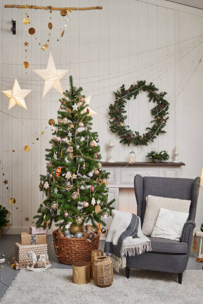istock room with fireplace and New Year tree. a spruce wreath hangs over the fireplace. the tree is decorated with gold and white toys. next to the tree there is a cozy gray armchair with a blanket on it 1331612757