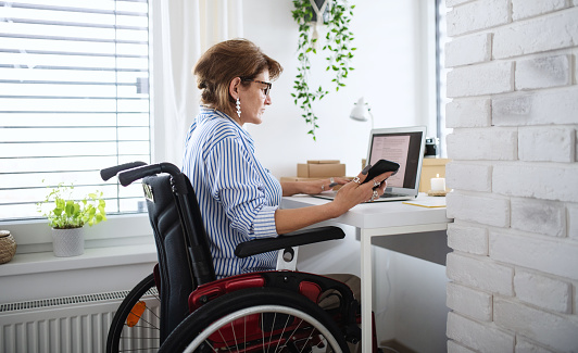A disabled mature woman with laptop working indoors in home office, small business concept.