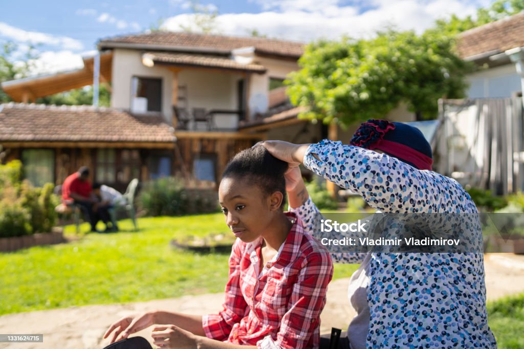 Hairstyling in the backyard Afro-American mom is brushing her daughters' hair while they are sitting outside in the sunshine of their backyard 12-13 Years Stock Photo