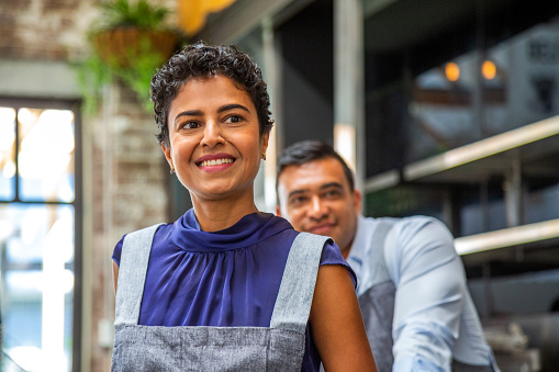 Happy, smiling couple of owners/co-workers in small cafe with woman in foreground and man in background (soft focus): small business, Pacific Islander, Indian and Latin American & Hispanic ethnicities