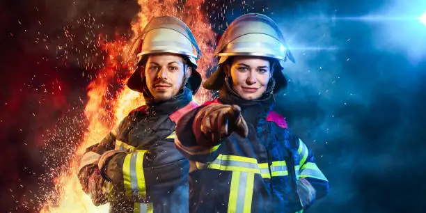 The two firefighters are standing in a dark scene in front of fire and blue light.The women is pointing with her finger through the camera.