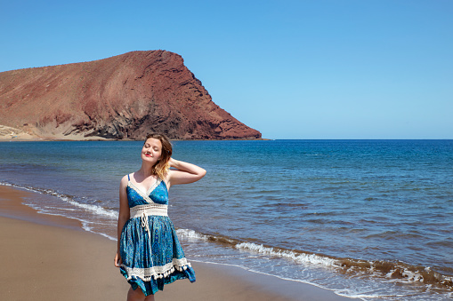 Beautiful millennial woman holding her hair with one hand and the head tilted with her eyes closed while enjoying Playa la Tejita, a volcanic beach with view to Montana Roja, Tenerife, Canary Islands, Spain.