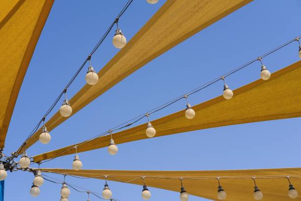 파란색 하늘 배경에 파티 밤 조명전구 램프의 행캔버스 항해로 만든 노란색 직물 스트레치 지붕의 동적 통풍이 잘되는 질감. - shade sail awning textile 뉴스 사진 이미지