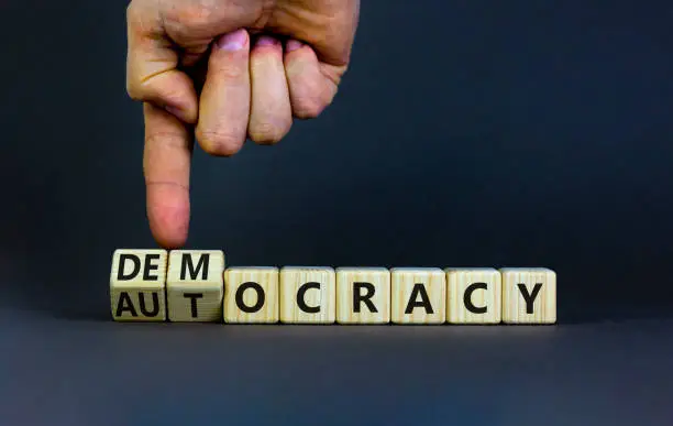 Photo of Democracy or autocracy symbol. Businessman turns wooden cubes and changes the word autocracy to democracy. Beautiful grey background, copy space. Business and democracy or autocracy concept.