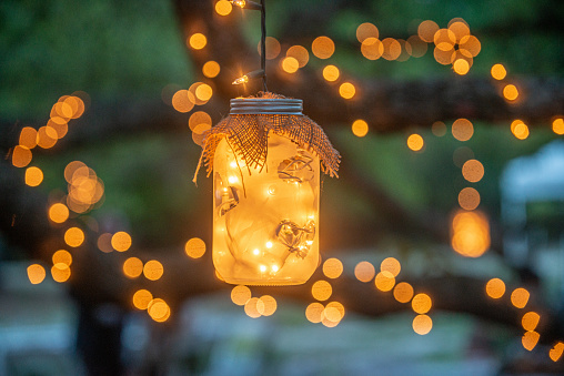 Mason jars filled with warm glowing lights decorations hanging from trees drapped in lights, close up