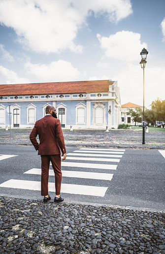 View from behind of a fashionable bald bearded guy in an elegant costume of a chestnut color and eyeglasses, ready to cross the road of a European city on the pedestrian crossing, Lisbon, Portugal