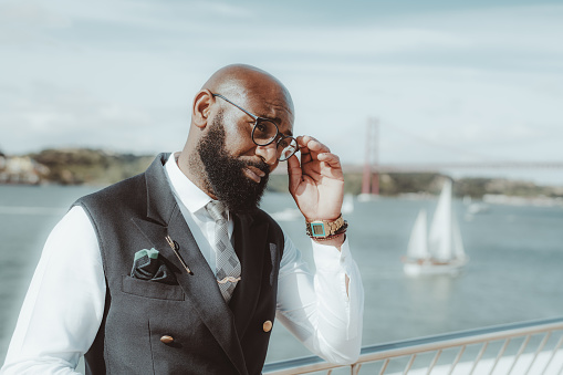 A dapper African American senior with a well-groomed black beard and in a fashionable formal suit with a vest is adjusting his elegant eyeglasses while standing on the pier with a sailboat behind him