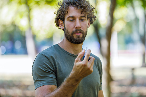 A Man with Breathing Problems is Feeling Displeased and Using a Nasal Spray During a Walk in City Park During a Summer Day.