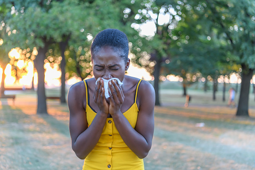 An African-American Woman with Sinusitis Problems is Feeling Displeased and Blowing Nose in Napkin During a Walk in City Park During a Summer Day.
