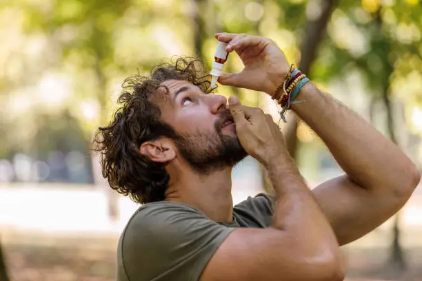 A Young Bearded Man is Walking in Public Park and Dropping Liquid in his Eye with Eye-dropper. Vision and Ophthalmology Medicine Health Concept.