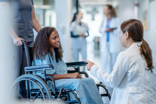 A teen girl with a disability sits in a wheelchair and smiles while talking to her doctor.